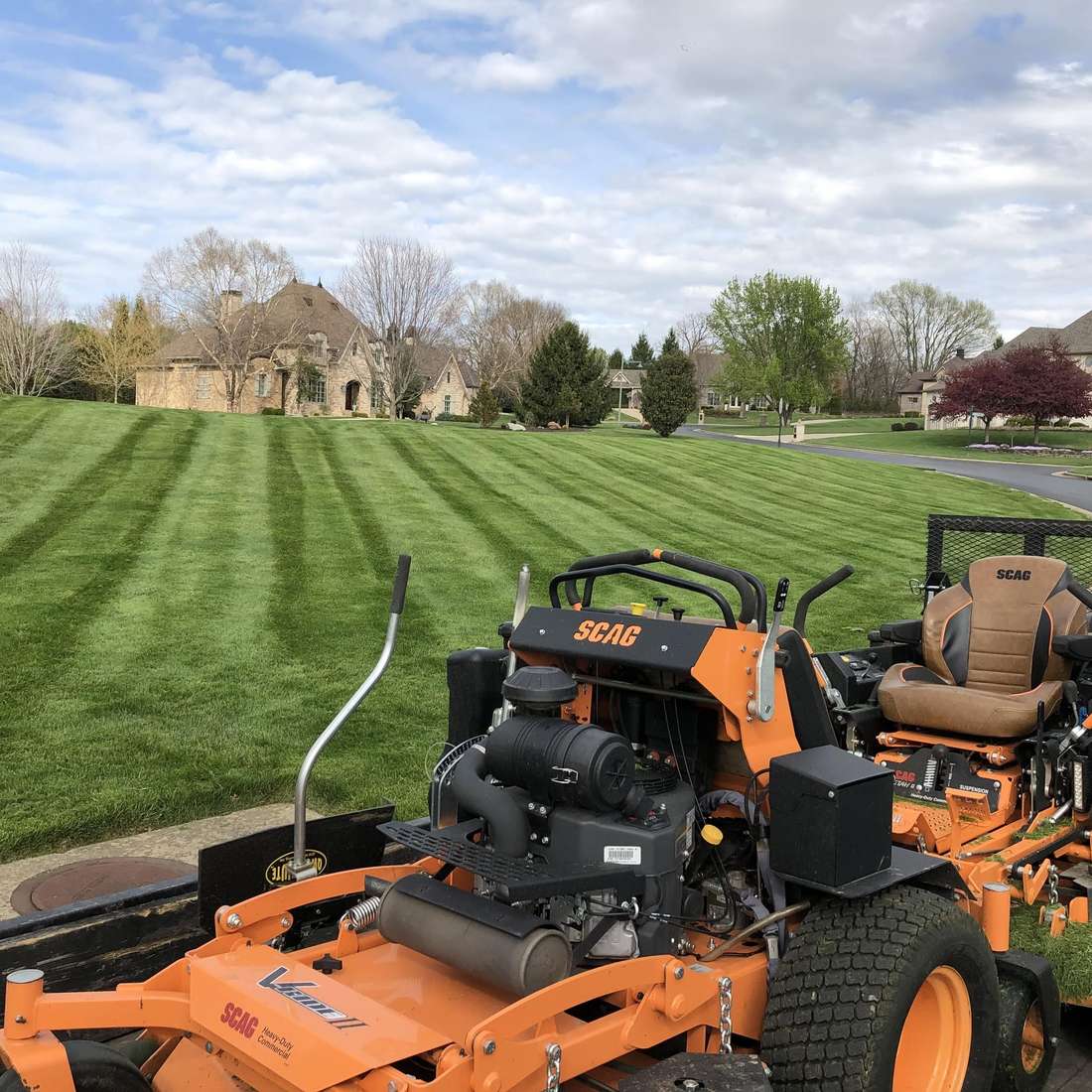 Two commercial zero-turn mowers on a trailer in front of a large freshly striped lawn and stone house